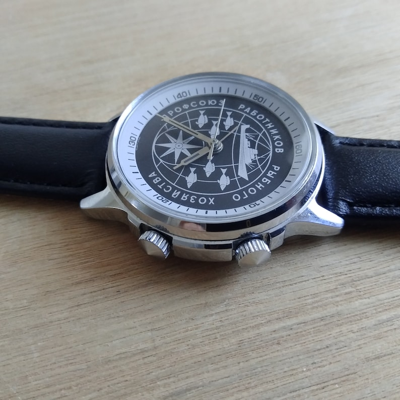 Puede incluir: Un reloj de pulsera plateado con una esfera negra y una correa de cuero negra. La esfera del reloj presenta un dise&ntilde;o de br&uacute;jula con el texto "ХОЗЯЙСТВА РАБОТНИКОВ РЫБОХОЛО  ПРОФСОЮЗ" alrededor del borde.