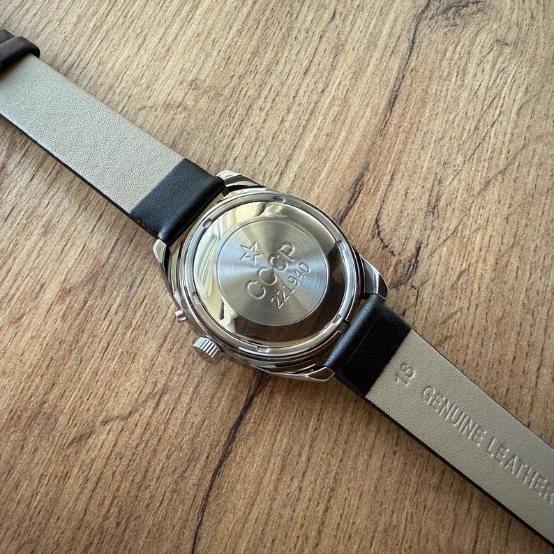 May include: A silver-toned wristwatch with a black leather band. The watch face is visible, displaying the text "CCCP" and "221940". The watch band is marked "18 GENUINE LEATHER". The watch is set against a wooden surface.