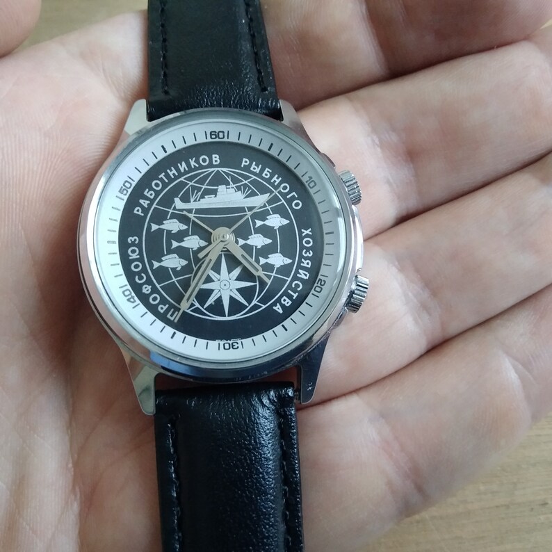 Puede incluir: Un reloj de pulsera plateado con una correa de cuero negro. La esfera del reloj tiene un fondo negro con una br&uacute;jula, un barco y peces. El texto en la esfera del reloj dice "ПРОФСОЮЗ РАБОТНИКОВ РЫБНОГО ХОЗЯЙСТВА".