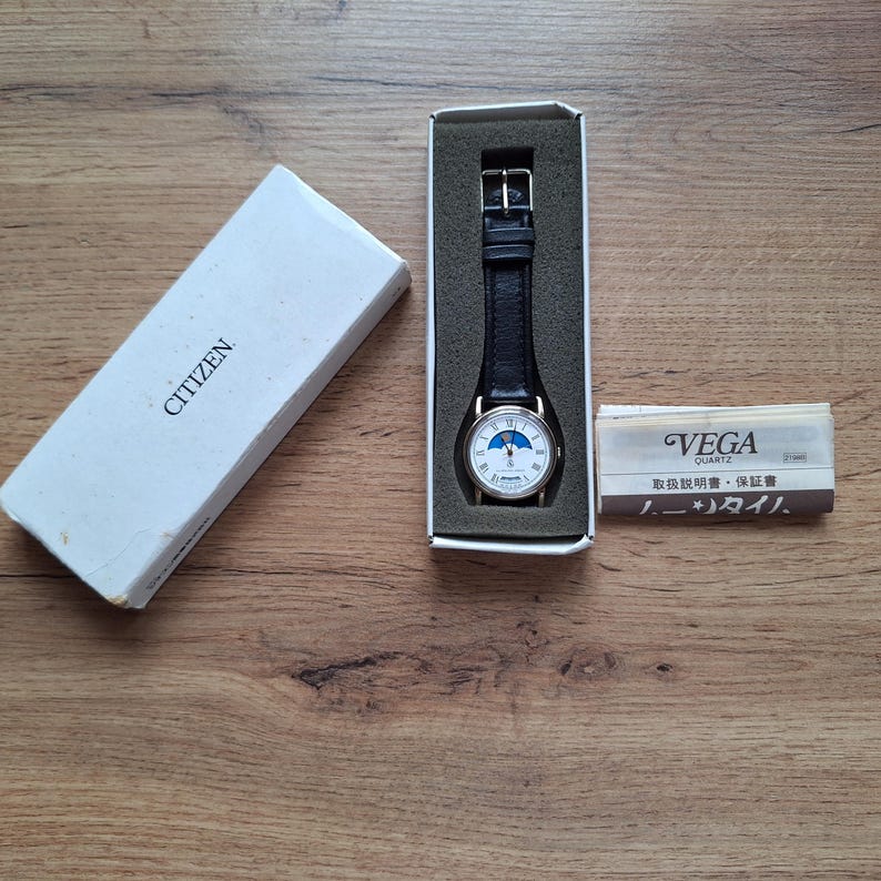 Op de afbeelding: Een Citizen quartz horloge met een zwarte leren band, gepresenteerd in de originele doos. De wijzerplaat is wit met blauwe en gouden accenten. De doos is wit met het Citizen-logo. Er is ook een kleine instructieboekje aanwezig.