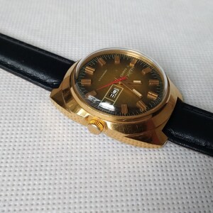 May include: A gold-tone wristwatch with a black leather band. The watch face is green with gold-tone hands and markers. The watch has a date window at the 3 o'clock position.
