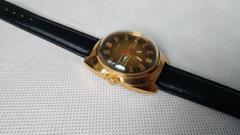 May include: A gold-tone wristwatch with a black leather band. The watch face is green with gold-tone hands and markers. The watch has a date window at the 3 o'clock position.