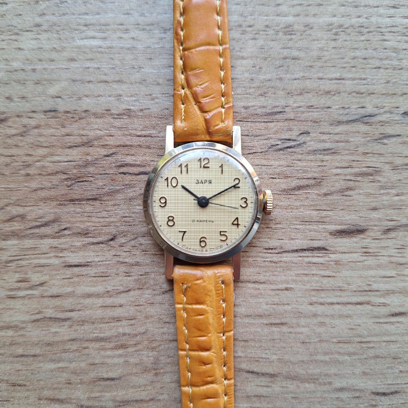 Op de afbeelding: Een vintage Zaria polshorloge met een lichtgekleurde wijzerplaat, zwarte cijfers en wijzers. Het horloge heeft een goudkleurige kast en een bruinleren band. De band is ongeveer 1,3 cm breed.