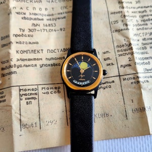 Pode incluir: Um relógio de pulso dourado com um mostrador preto e uma pulseira de couro preta. O mostrador do relógio apresenta uma exibição de fase lunar e uma janela de data. O relógio está em um fundo branco com texto em russo.