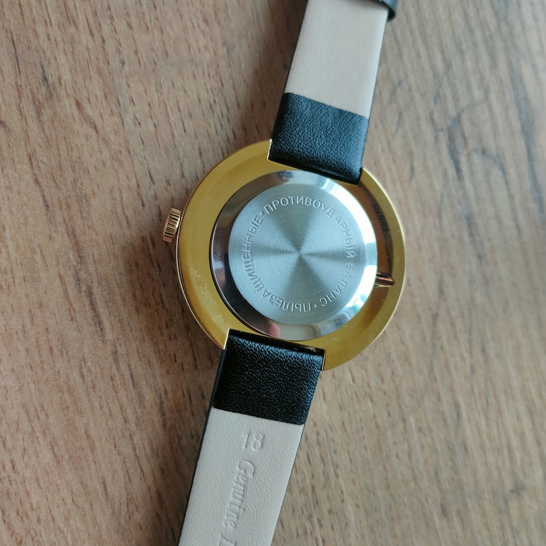 May include: A gold-tone wristwatch with a black leather band. The back of the watch has a silver-tone case with the text "ЧНЦУ. РИПЛЕБА. ПРОТИВОУДАРНЫЕ" in Cyrillic.
