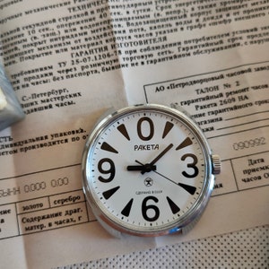 May include: A white-faced Raketa watch with black numerals 0, 3, 6, and 9. The watch has a silver bezel and black hands. The dial includes the word "Raketa" and a star symbol. The watch rests on a paper document.