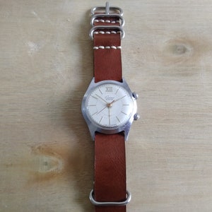 Op de afbeelding: Een zilveren polshorloge met een witte wijzerplaat en zwarte wijzers. Het horloge heeft een bruin leren bandje met witte stiksels en zilveren gespen. De wijzerplaat van het horloge heeft de tekst "Cucuruz" in gouden letters.