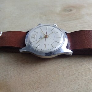Op de afbeelding: Een zilveren polshorloge met een witte wijzerplaat en zwarte Romeinse cijfers. Het horloge heeft een bruin leren bandje en de woorden "Guintex Automatic" staan op de wijzerplaat gedrukt.