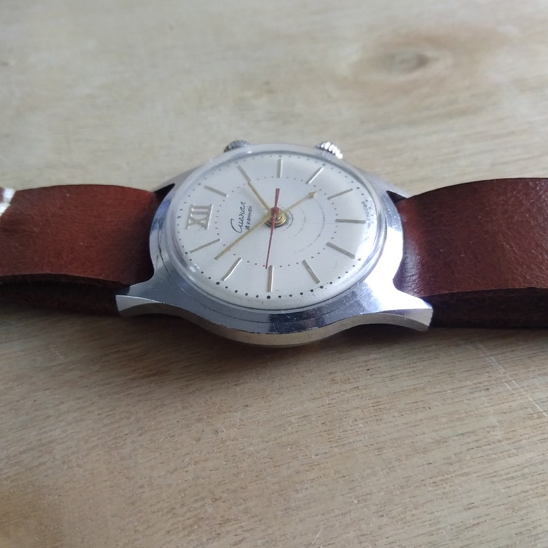 Op de afbeelding: Een zilveren polshorloge met een witte wijzerplaat en zwarte Romeinse cijfers. Het horloge heeft een bruin leren bandje en de woorden "Guintex Automatic" staan op de wijzerplaat gedrukt.