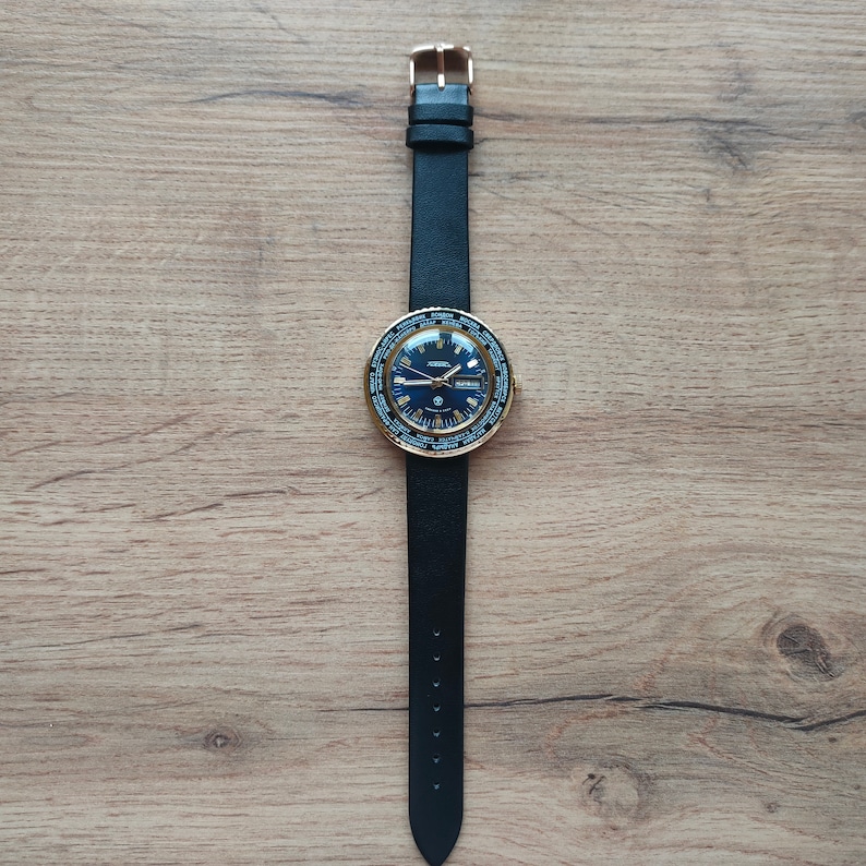 May include: A gold-tone wristwatch with a black leather band and a blue face. The watch has a world time function and a date window.