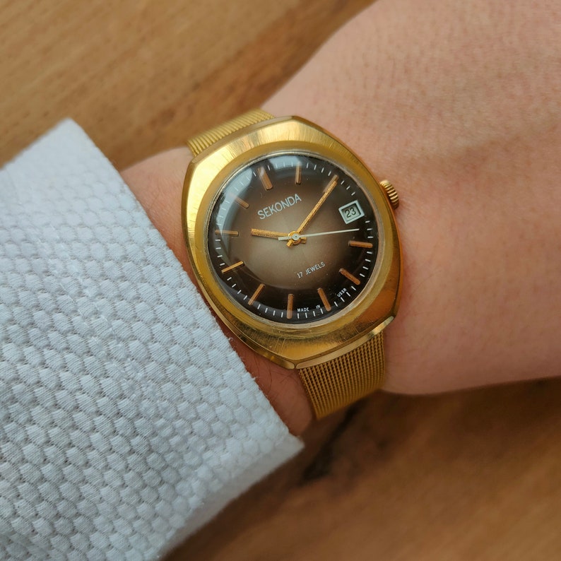 Puede incluir: Un reloj Sekonda dorado con esfera degradada marr&oacute;n y visualizaci&oacute;n de fecha. El reloj tiene una correa de malla y est&aacute; marcado "17 Jewels". El reloj est&aacute; en una mu&ntilde;eca, junto a una camisa blanca texturizada.