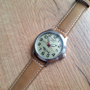 K&ouml;nnte beinhalten: Eine braune Lederarmbanduhr mit einem silbernen Geh&auml;use und einem wei&szlig;en Zifferblatt. Das Zifferblatt hat schwarze Zahlen und Zeiger, einen roten Sekundenzeiger und eine kleine Flugzeuggrafik. Auf dem Zifferblatt der Uhr steht auch "Aviator" und "Lechtad N. 1952-2022".