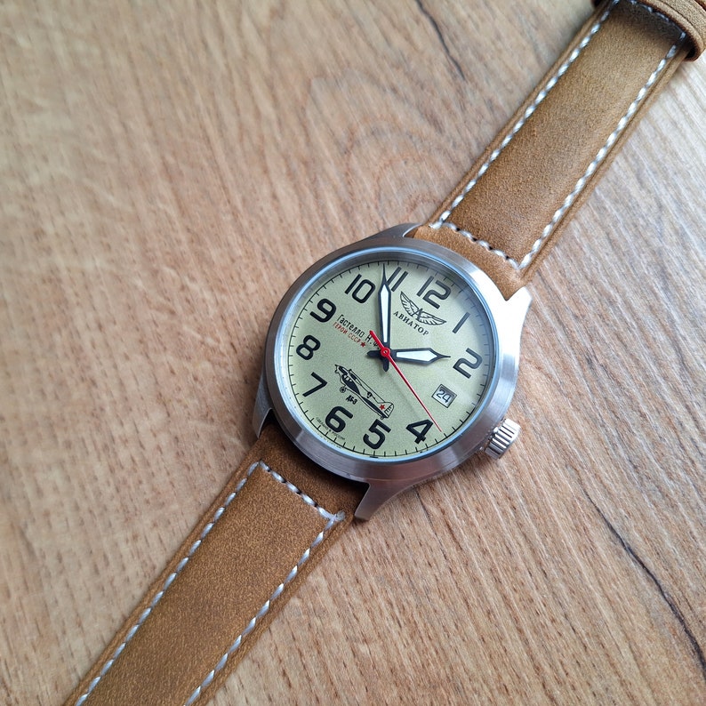 K&ouml;nnte beinhalten: Eine braune Lederarmbanduhr mit einem silbernen Geh&auml;use und einem wei&szlig;en Zifferblatt. Das Zifferblatt hat schwarze Zahlen und Zeiger, einen roten Sekundenzeiger und eine kleine Flugzeuggrafik. Auf dem Zifferblatt der Uhr steht auch "Aviator" und "Lechtad N. 1952-2022".