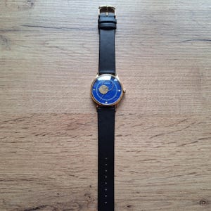 Op de afbeelding: Een gouden polshorloge met een blauwe wijzerplaat en een zwart leren bandje. Het horloge heeft een uniek ontwerp met een ronde uitsparing in het midden van de wijzerplaat.