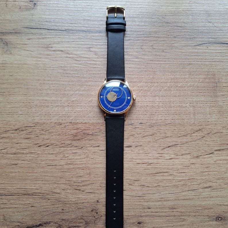 Op de afbeelding: Een gouden polshorloge met een blauwe wijzerplaat en een zwart leren bandje. Het horloge heeft een uniek ontwerp met een ronde uitsparing in het midden van de wijzerplaat.