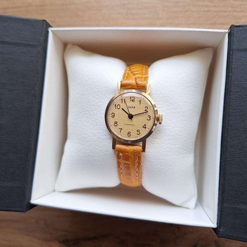 Op de afbeelding: Een vintage goudkleurig polshorloge met een lichtbruine leren band, gepresenteerd in een witte doos. De wijzerplaat heeft zwarte cijfers en wijzers, met het woord "SAPR" boven het midden gedrukt.