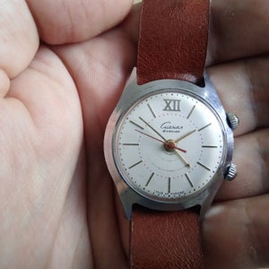 Op de afbeelding: Een zilveren polshorloge met een witte wijzerplaat en zwarte wijzers. Het horloge heeft een bruin leren bandje en staat "Cиeзxaл 18 камней" op de wijzerplaat.