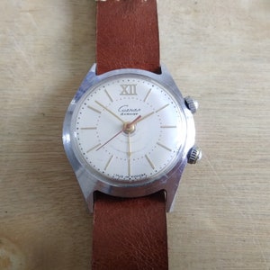 Op de afbeelding: Een zilveren polshorloge met een witte wijzerplaat en zwarte wijzers. Het horloge heeft een bruin leren bandje en staat "Слава 18 камней" op de wijzerplaat.