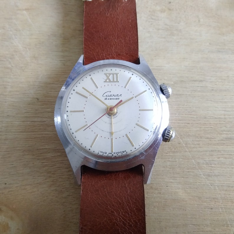Op de afbeelding: Een zilveren polshorloge met een witte wijzerplaat en zwarte wijzers. Het horloge heeft een bruin leren bandje en staat "Слава 18 камней" op de wijzerplaat.