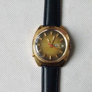 May include: A gold-tone wristwatch with a black leather band. The watch face is green with gold-tone markings and a date window. The watch face says "Чайка" in Cyrillic letters.