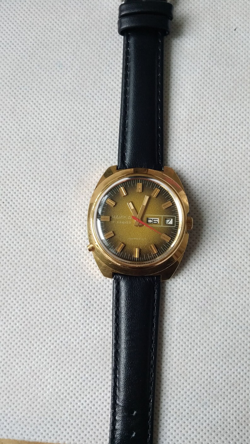 May include: A gold-tone wristwatch with a black leather band. The watch face is green with gold-tone markings and a date window. The watch face says "Чайка" in Cyrillic letters.