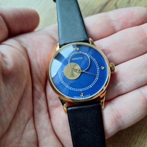 Op de afbeelding: Een gouden polshorloge met een blauwe wijzerplaat en een zwart leren bandje. De wijzerplaat van het horloge heeft een gouden maanfase-indicator en de tekst "PAKEТА" in cyrillisch schrift.