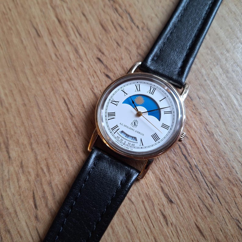 Op de afbeelding: Een goudkleurig polshorloge met een witte wijzerplaat met Romeinse cijfers en een maanfase-indicator. Het horloge heeft een zwarte leren band. De tekst "A.G. Spalding & Bros" is zichtbaar op de wijzerplaat. Op een houten achtergrond.