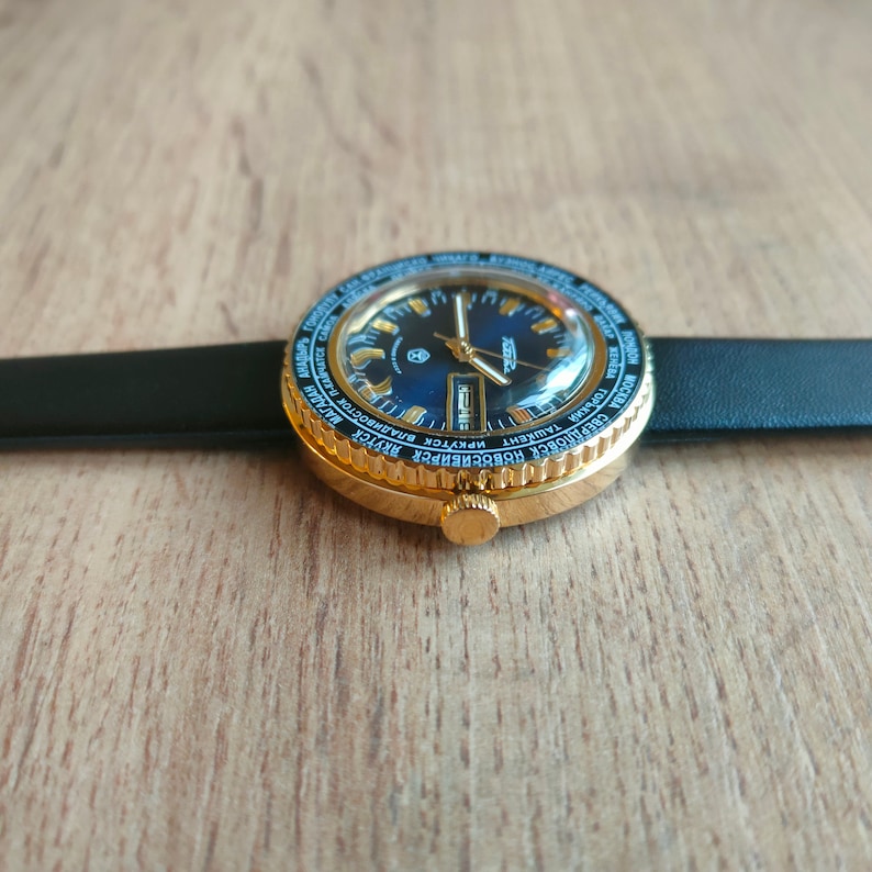 May include: A gold-toned wristwatch with a black leather band and a blue face. The watch face has a rotating bezel with city names printed around the edge.