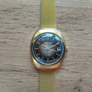 Puede incluir: Reloj de pulsera Sekonda dorado con correa de malla. La esfera marr&oacute;n tiene agujas e indicadores dorados, y una ventana de fecha. El texto "SEKONDA" es visible. El reloj est&aacute; sobre un fondo de madera.
