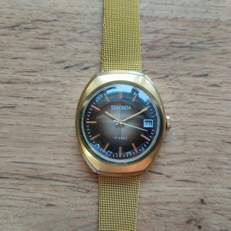 Puede incluir: Reloj de pulsera Sekonda dorado con correa de malla. La esfera marr&oacute;n tiene agujas e indicadores dorados, y una ventana de fecha. El texto "SEKONDA" es visible. El reloj est&aacute; sobre un fondo de madera.
