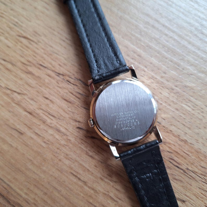 Op de afbeelding: Close-up van een goudkleurig Citizen horloge met een zwarte leren band. De wijzerplaat is zilverkleurig en de achterkant van het horloge heeft de tekst "Citizen Watch Co. Water Resist Base Metal YP 0196-408746 L 80500253 Japan-L12."