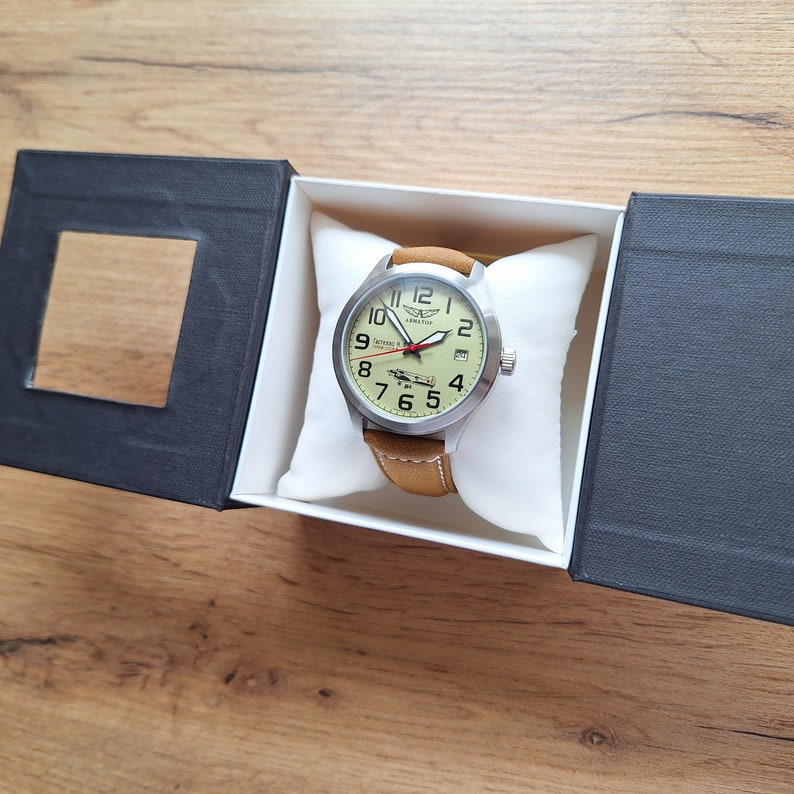 K&ouml;nnte beinhalten: Eine silberne Uhr mit einem braunen Lederarmband. Das Zifferblatt der Uhr ist wei&szlig; mit schwarzen Zahlen und einer kleinen Flugzeuggrafik. Die Uhr befindet sich in einer schwarzen Box mit einem wei&szlig;en Kissen.