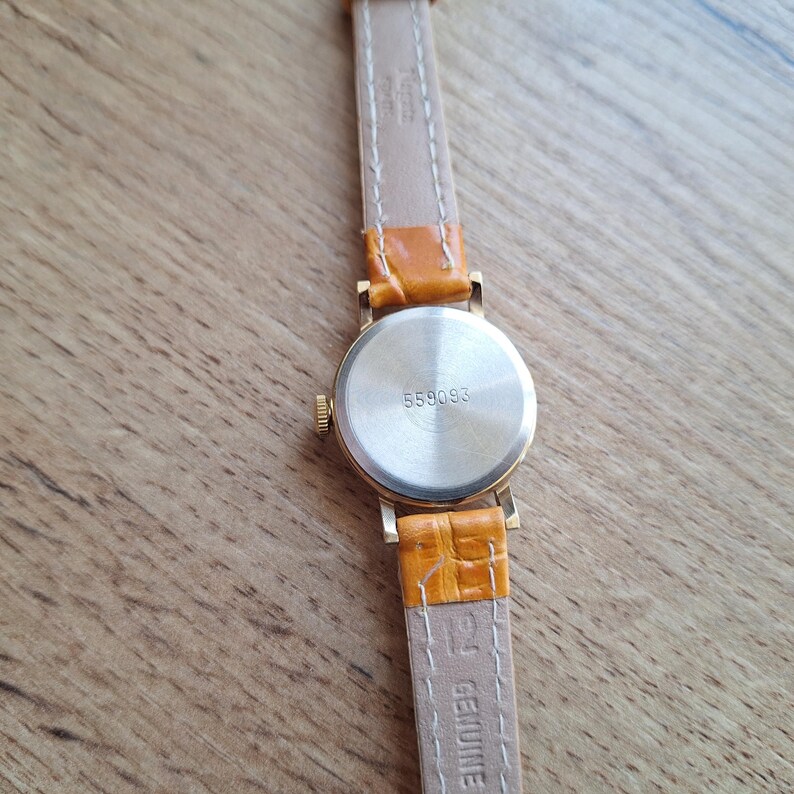 Op de afbeelding: Een goudkleurig polshorloge met een lichtbruine leren band. De wijzerplaat van het horloge is rond en de achterkant van het horloge heeft het nummer 558093. De horlogeband is lichtbruin met een getextureerde, oranje leren band.
