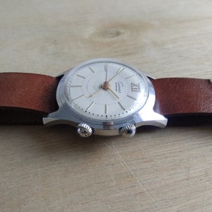 Op de afbeelding: Een zilveren polshorloge met een witte wijzerplaat en zwarte wijzers. Het horloge heeft een bruin leren bandje en een datumvenster op de 3 uur positie. Op de wijzerplaat van het horloge staat "Cucinat" en "17 Jewels" gedrukt.