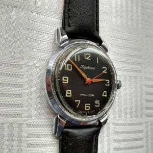 Pu&ograve; includere: Un orologio da polso argentato con quadrante nero e cinturino in pelle nera. Il quadrante dell'orologio ha numeri bianchi e una lancetta dei secondi rossa. Sul quadrante dell'orologio &egrave; anche stampato il testo "Kirovskie".