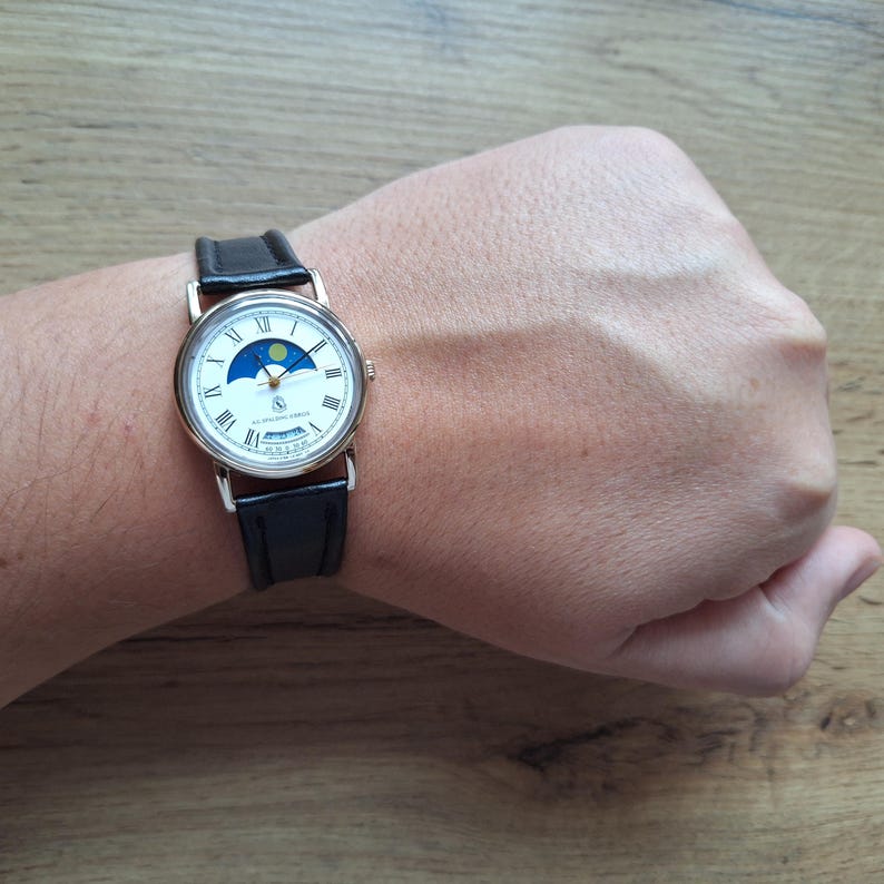 Op de afbeelding: Een goudkleurig polshorloge met een witte wijzerplaat, zwarte leren band en Romeinse cijfers. De wijzerplaat van het horloge heeft een maanfase-indicator en een datumweergave. Het horloge wordt om de pols gedragen.