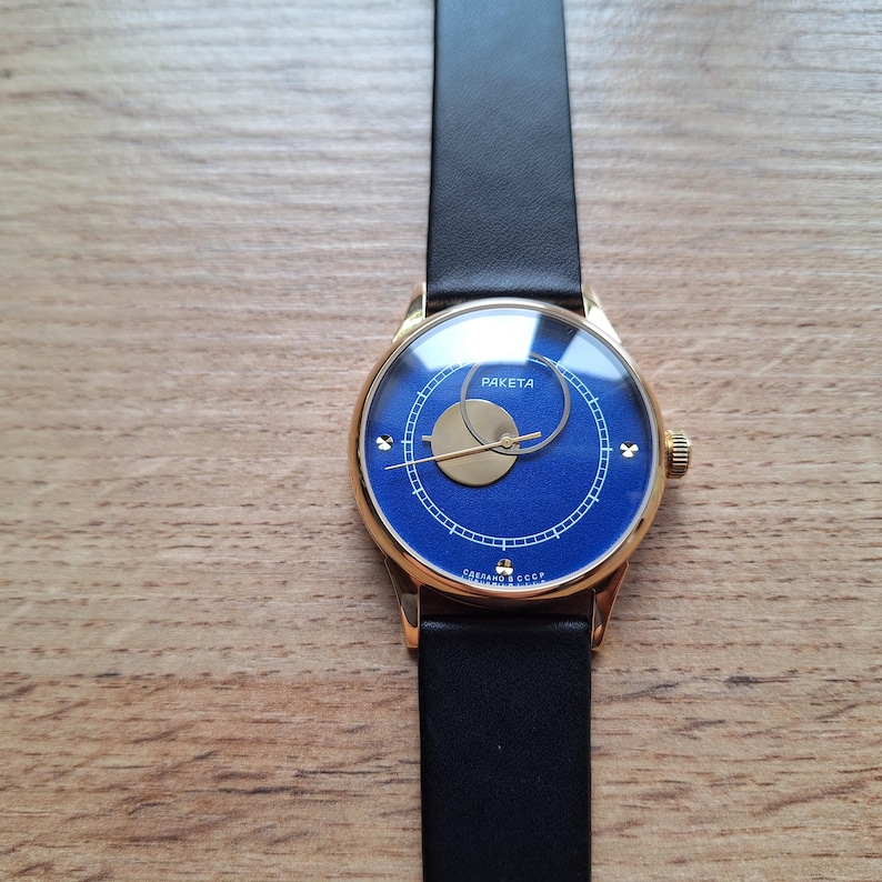 Op de afbeelding: Een gouden polshorloge met een blauwe wijzerplaat en een zwart leren bandje. De wijzerplaat van het horloge heeft een cirkelvormig ontwerp met het woord "PAKEta" en de tekst "СССР" in het Cyrillisch.