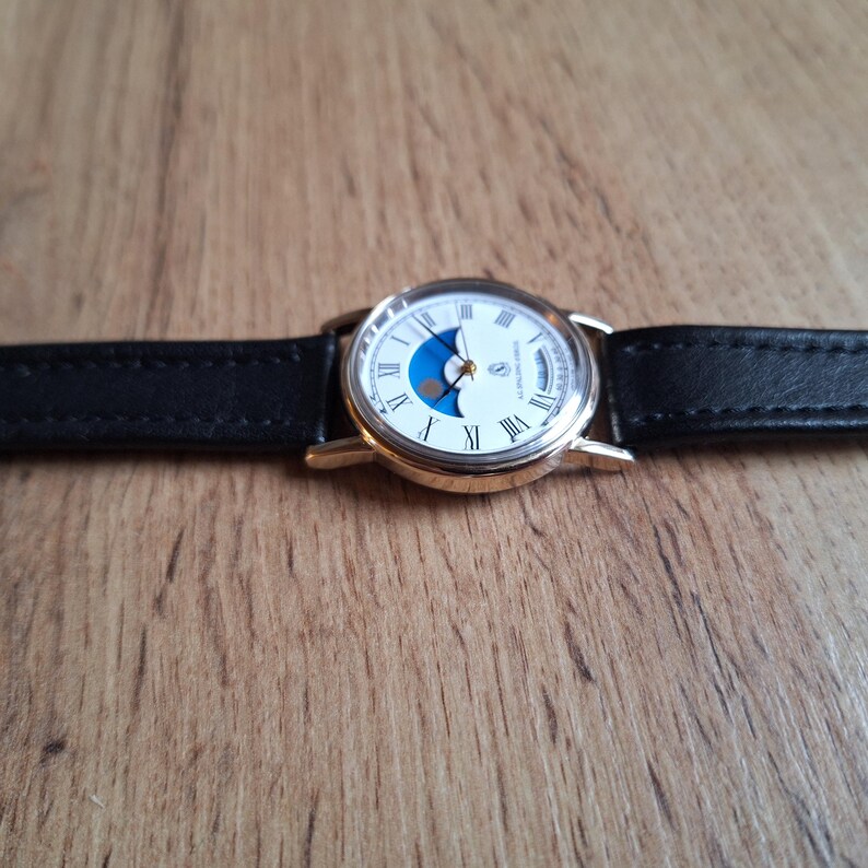 Op de afbeelding: Een goudkleurig polshorloge met een witte wijzerplaat, Romeinse cijfers en een blauw maansikkelontwerp. Het horloge heeft een zwarte leren band. Het horloge ligt op een houten oppervlak.