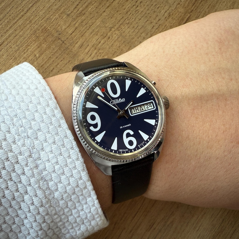 May include: A vintage wristwatch with a dark blue face, large white numerals, and a black leather band. The watch has a silver-colored bezel and a day-date display. The watch is on a wrist.