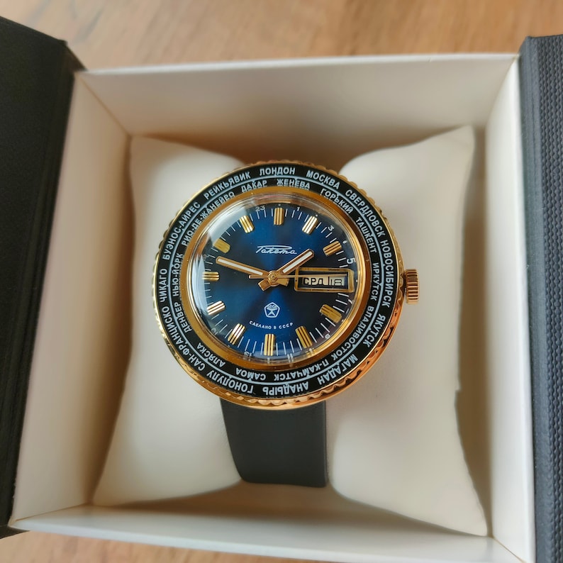 May include: A gold-toned wristwatch with a blue face and a black leather band. The watch face has a world map printed around the edge and a date window at the 3 o'clock position. The watch face also has the text "Слава" printed on it.