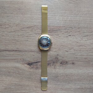 Puede incluir: Un reloj Sekonda dorado con esfera marr&oacute;n oscuro y correa de malla. El reloj tiene una esfera rectangular con visualizaci&oacute;n de fecha. La correa se asegura con un cierre. El reloj se muestra sobre una superficie de madera.