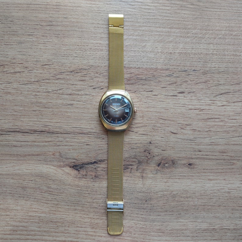 Puede incluir: Un reloj Sekonda dorado con esfera marr&oacute;n oscuro y correa de malla. El reloj tiene una esfera rectangular con visualizaci&oacute;n de fecha. La correa se asegura con un cierre. El reloj se muestra sobre una superficie de madera.