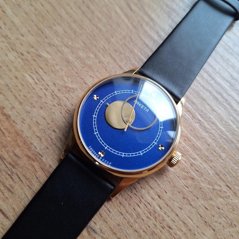 Op de afbeelding: Een gouden polshorloge met een blauwe wijzerplaat en een zwarte leren band. De wijzerplaat van het horloge heeft een witte wijzerplaat met zwarte markeringen en het woord "RAKETa" erop gedrukt. Het horloge heeft ook een klein, rond venster op de 12-uur positie.