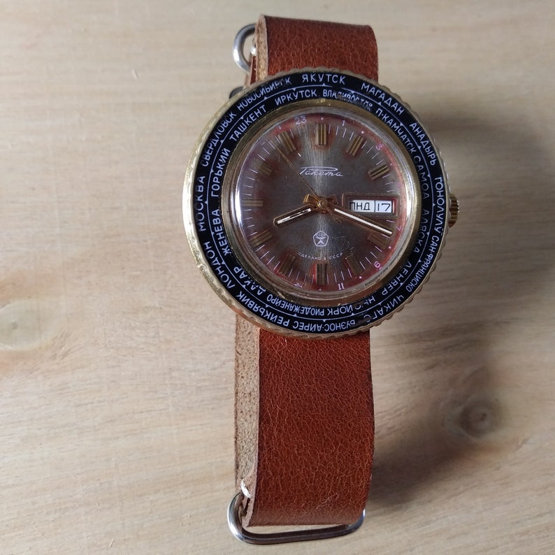 Puede incluir: Un reloj de pulsera dorado con una correa de cuero marr&oacute;n. La esfera del reloj tiene un anillo exterior negro con nombres de ciudades impresos en negro. La esfera del reloj es plateada con marcadores de hora romanos negros y agujas negras. La ventana de fecha est&aacute; en la posici&oacute;n de las 3 en punto y muestra la fecha "ПНД 17".