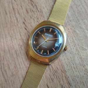 Puede incluir: Un reloj de pulsera Sekonda dorado con una esfera degradada marr&oacute;n y dorada. El reloj tiene una correa de malla y una pantalla de fecha a las 3 en punto. La esfera del reloj dice "Sekonda" y "17 Jewels".