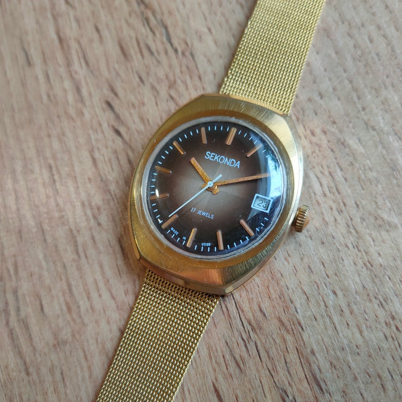 Puede incluir: Un reloj de pulsera Sekonda dorado con una esfera degradada marr&oacute;n y dorada. El reloj tiene una correa de malla y una pantalla de fecha a las 3 en punto. La esfera del reloj dice "Sekonda" y "17 Jewels".