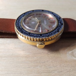 Puede incluir: Un reloj de pulsera dorado con un bisel negro y una esfera de hora mundial. La esfera del reloj es plateada con marcadores de hora romanos negros y una ventana de fecha. La correa del reloj es de cuero marr&oacute;n.