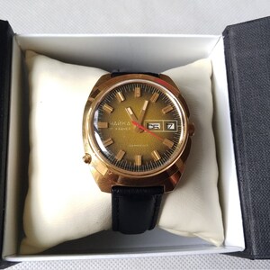 May include: A gold-tone wristwatch with a black leather band. The watch face is a gradient of green and gold with black markings and a date window. The watch is in a white box with a black interior.