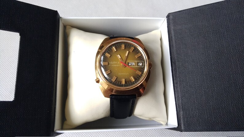 May include: A gold-tone wristwatch with a black leather band. The watch face is a gradient of green and gold with black markings and a date window. The watch is in a white box with a black interior.