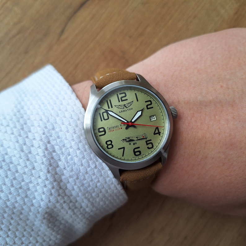 K&ouml;nnte beinhalten: Eine silberne Armbanduhr mit einem braunen Lederarmband. Das Zifferblatt der Uhr ist gr&uuml;n mit wei&szlig;en Zahlen und Zeigern. Die Uhr hat eine kleine Flugzeuggrafik und den Text "Гастелло Н. И. 17.04.1942" auf dem Zifferblatt. Die Marke der Uhr ist "ARMATOF".